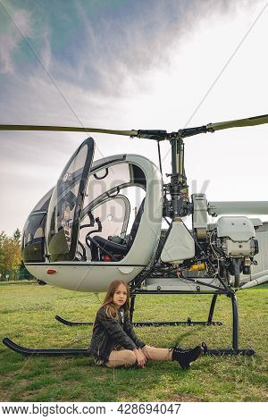 Tween Girl Sitting On Green Grass Of Flying Field Near Helicopter