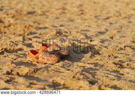 A Pair Of Children's Orange Beach Sandals On Yellow Sand. Nobody. The Concept Of Recreation And Chil