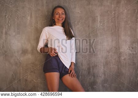 Slender Joyful Sporty Brunette Woman In Sports Shorts And Jumper Standing Leaning On Concrete Wall I