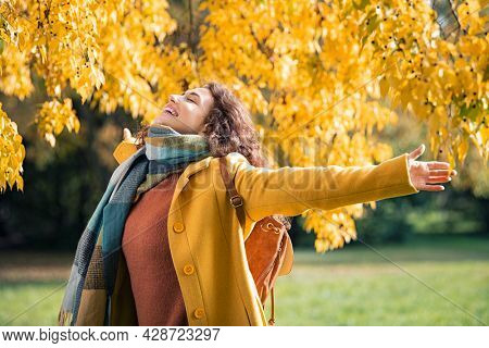 Beautiful young woman relaxing at park during autumn season with closed eyes. Happy free natural girl breathing deeply in park with foliage in background. Pretty woman expressing freedom outdoor.