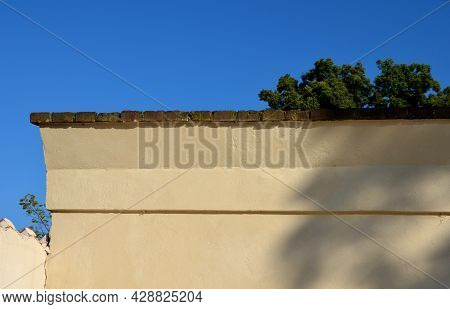 Crown Wall, Garden Fence. Fired Bricks Protect The Low Thick Plastered Wall Of Yellow Color. Wall In