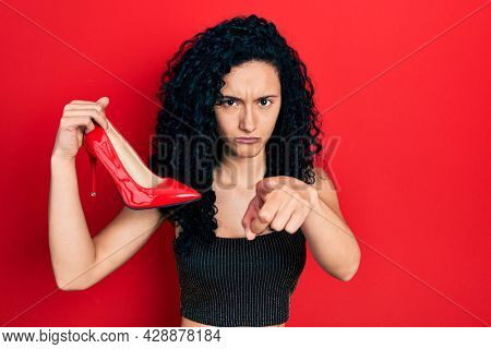 Young hispanic woman with curly hair holding red high heel shoes pointing with finger to the camera and to you, confident gesture looking serious 