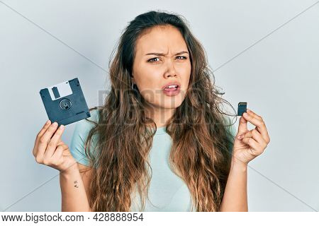 Young hispanic girl holding floppy disk and sdxc card clueless and confused expression. doubt concept. 