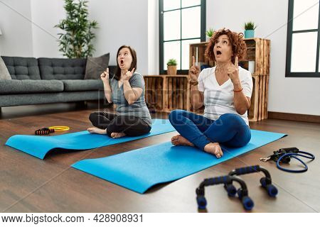 Family of mother and down syndrome daughter doing exercise at home amazed and surprised looking up and pointing with fingers and raised arms. 