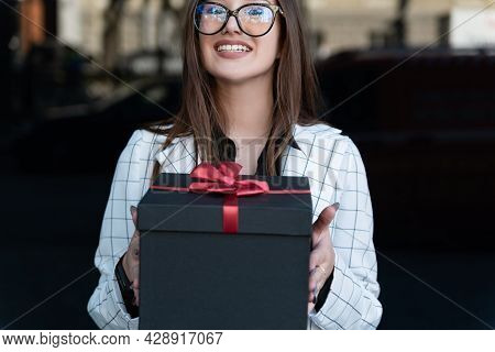 Young Stylish Woman Hands Gift. Happy Young Woman Holds Gift Box And Smiling. Black Gift Box With Re