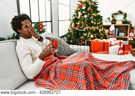 Young african american man sitting on the sofa drinking coffee by christmas tree smelling something stinky and disgusting, intolerable smell, holding breath with fingers on nose. bad smell 