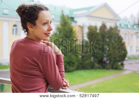A young woman is standing on the balcony and watching outside leaning on railing
