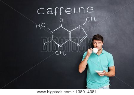 Handsome young man drinking coffee over blackboard background with drawn chemical structure of caffeine molecule