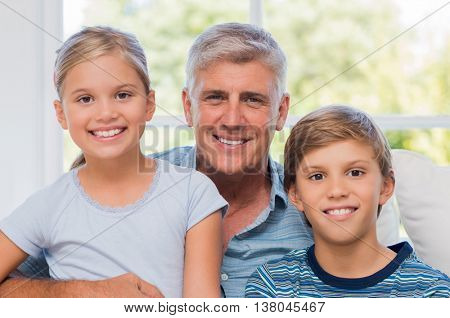 Grandpa sitting with boy and girl looking at camera. Cheerful Grandfather embracing grandson and granddaughter on couch. Portrait of smiling grandchildren with older man.