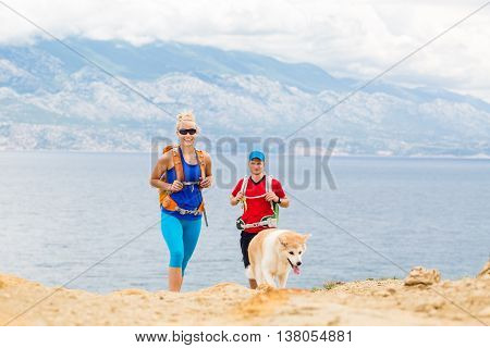 Happy couple hikers trekking with akita dog in summer mountains at seaside. Young woman and man walking on rocky mountain trail path looking at beautiful inspirational landscape sea view.