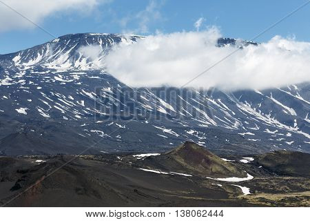 Volcanic landscape of Kamchatka Peninsula: beautiful summer view of cone of active Plosky Tolbachik Volcano and clouds partially hiding the top of volcano. Eurasia Russian Far East Kamchatsky Region Klyuchevskaya Group of Volcanoes.