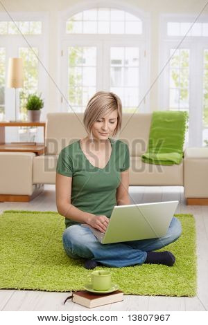 微笑的女人坐在客厅里为远程办公使用便携式计算机在家里地板上。?