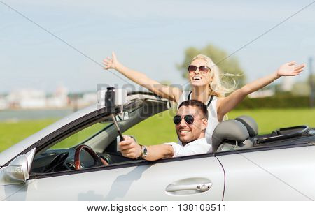 road trip, leisure, couple, technology and people concept - happy man and woman driving in cabriolet car and taking picture with smartphone on selfie stick
