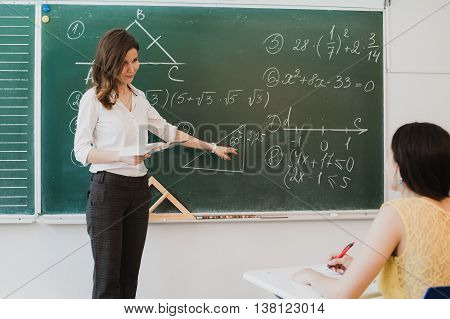 Teacher or docent or educator giving while lesson in front of a blackboard or board a sheet of paper and educate or teaching students or pupils or mates in a school or class.