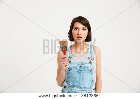 A portrait of young pretty girl, in gray shirt and denim overall,  holding painting brush, isolated on white background