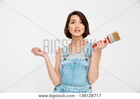 A portrait of young pretty girl, in gray shirt and denim overall, looking amazed, holding painting brush, isolated on white background