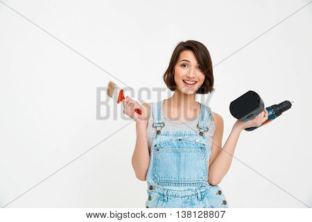 A portrait of young smiling  beautiful girl, in gray shirt and denim overall, holding painting brush and drill, looking at camera, isolated on white background