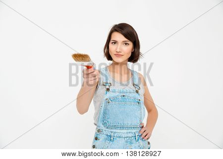 A portrait of young  beautiful girl, in gray shirt and denim overall, pointing forward with painting brush, looking at camera, isolated on white background