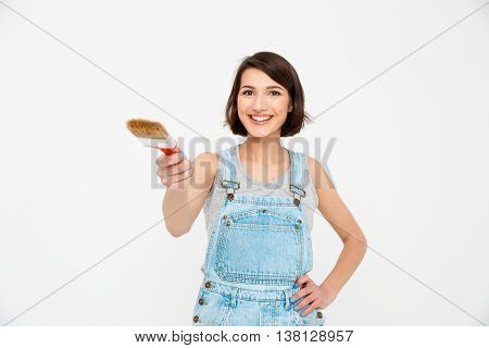 A portrait of young  beautiful smiling girl, in gray shirt and denim overall, pointing forward with painting brush, looking at camera, isolated on white background