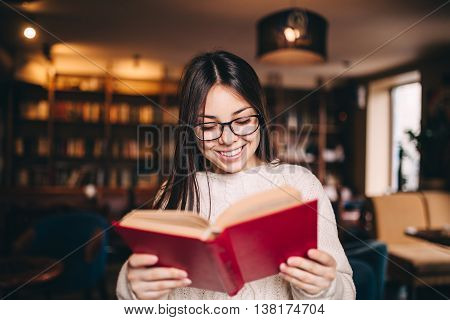 Smiling student girl in eyeglasses reading a red book in a library. School and education concept