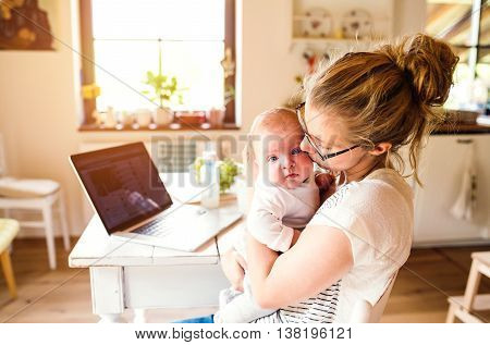 Close up of beautiful mother holding her baby son in the arms, kissing him, laptop and milk in the bottle laid on table