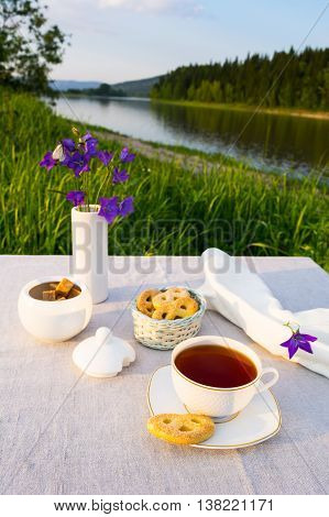 Evening tea sunset river view. Cup of tea and cookies in small wicker basket with blue bell flowers in vase. Summer tea time concept.