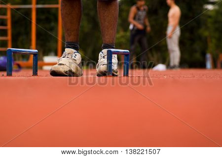 White shoes with mans legs standing on orange athletic running surface between two florr mounted blue handles, blurry people training background.