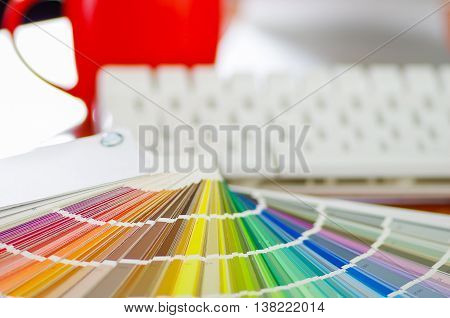 Closeup large palette colormap spread out in front of white computer keyboard and red coffee mug sitting on wooden desk, designer concept.