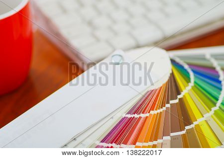 Closeup large palette colormap spread out in front of white computer keyboard and red coffee mug sitting on wooden desk, designer concept.