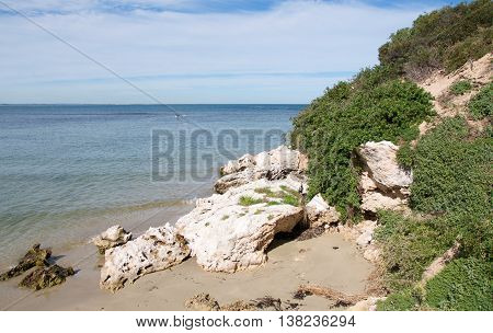 Coastal dunes with native plants, limestone rock and Indian Ocean waters under a blue sky with clouds in Rockingham, Western Australia.