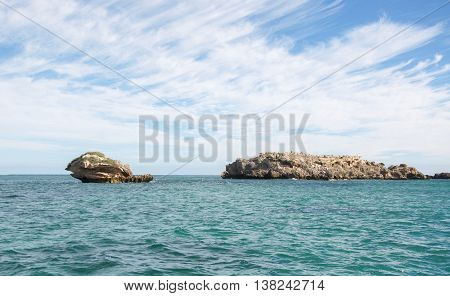 Limestone rock formations off the coast of Rockingham, Western Australia in the turquoise Indian Ocean waters under a blue sky with clouds.