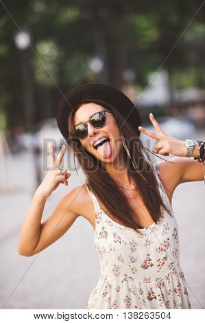 Gorgeous Fashion Funky Girl wearing sunglasses and hat swowing tongue and peace sign outdoors on a summer day