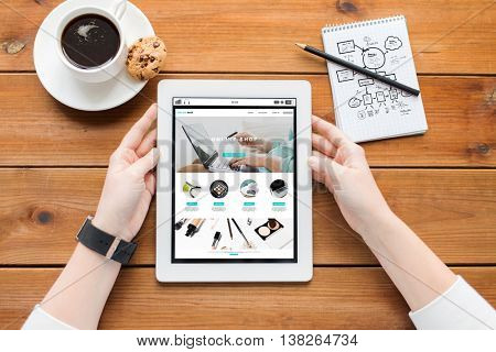 business, internet shopping, technology and people concept - close up of woman with online shop web page on tablet pc computer screen, notebook and coffee on wooden table