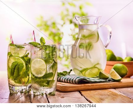 Country drink. On wooden boards is glass jug with transparent drink and lime glue bowl. Drink number hundred fifty with ice cubes on checkered napkin. Country life. Light background.
