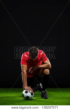 Soccer player in black and red uniform with ball standing on one knee over black background with head down