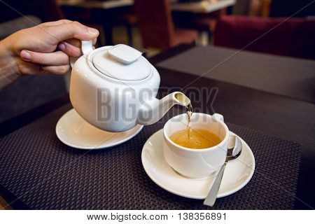 man pours tea from a white teapot in a tea Cup, saucer and teaspoon, a sugar bowl are on the table