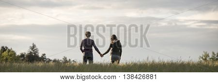 Wide panoramic view on couple holding hands in field from behind below partly cloudy sky with copy space.