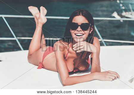 Enjoying this summer. Cheerful young woman in red bikini holding hand on chin and smiling at camera while lying on the deck of yacht