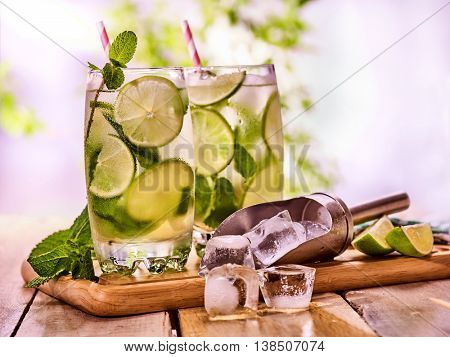 Alcohol drink. On wooden boards two glasses with alcohol drink and ice cubes. A drink number hundred fifty eight cocktail mohito and scoop ice with straw. Country still life outdoor. Light background.