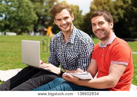 Before exam. Positive young man using a laptop and another delighted young man making some notes while being in the park and sitting on the grass together