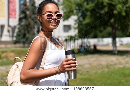 Just smile. Happy and delighted young woman holding thermos and smiling at a camera walking in the park