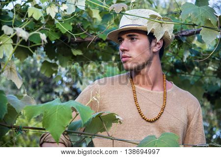 Side view of handsome young man in hat toching vine leaves in garden in sunlight