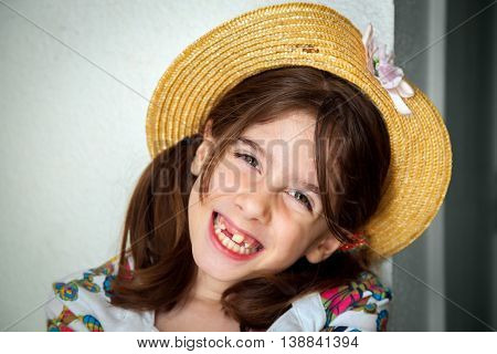 A cute young snaggletooth girl smiles. She is wearing a straw hat and is tilting her head to the side.
