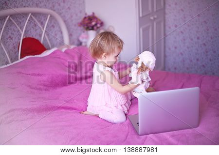 baby in pink room sitting on the bed and playing or watching cartoons price. Girl sitting at home. little princess in dress uses advanced fashion technology