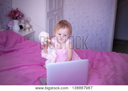 baby in pink room sitting on the bed and playing or watching cartoons price. Girl sitting at home. little princess in dress uses advanced fashion technology