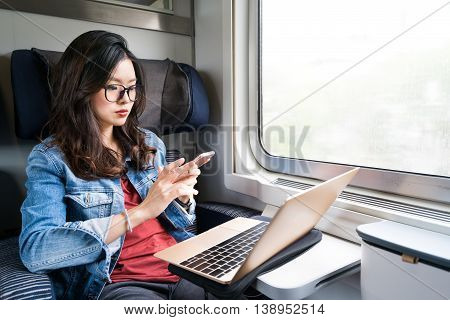 Cute Asian woman using smartphone and laptop on train copy space on window business travel or technology concept