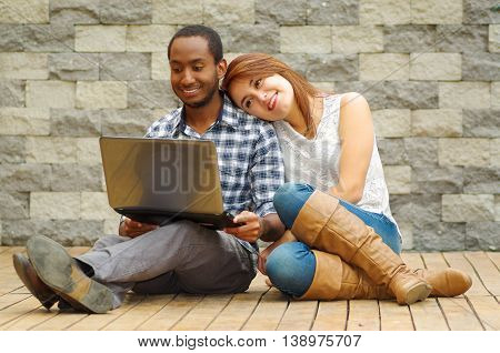 Interracial charming couple wearing casual clothes sitting down on wooden surface lookin at laptop together, in front of grey brick wall.