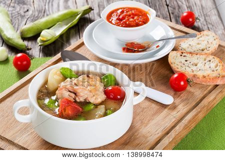 Braised chicken things with new potatoes tomatoes and butter lima bean in graten dish and in soup bowl on table mat tomato and bell pepper sauce in gravy boat view from above
