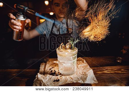 Barman Prepares Cocktail With Orange And Herbs In Transparent Glass On Bar With Alcohol. Uses Burner