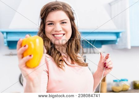 Portrait Of Smiling Overweight Woman In Headphones Showing Fresh Bell Pepper In Hand In Kitchen